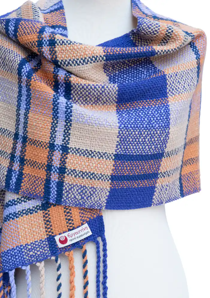 A custom-made handmade woven scarf by Ergastini, featuring a beautiful blue, peach, and cream plaid pattern with twisted fringes, draped on a mannequin.