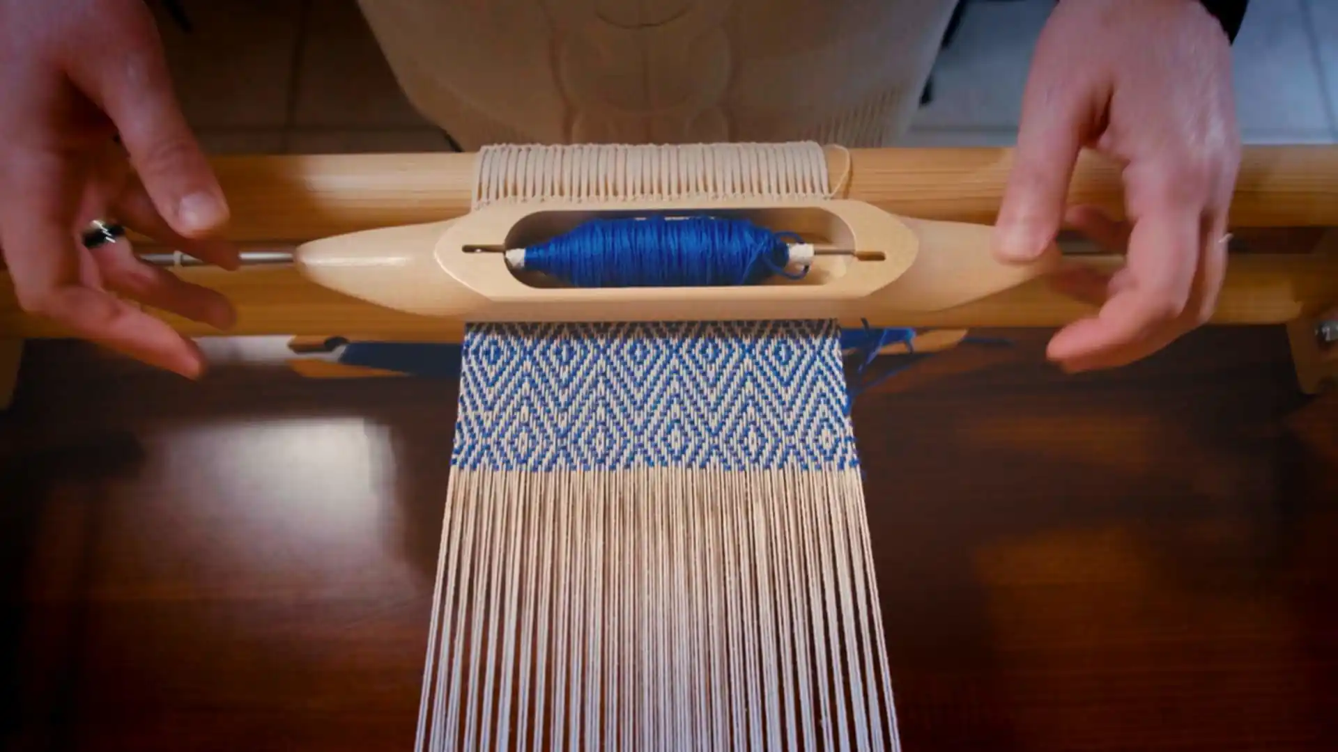 Zoe's hands holding a wooden weaving shuttle with bright blue yarn over a loom, actively weaving a complex blue and white geometric pattern for Ergastini
