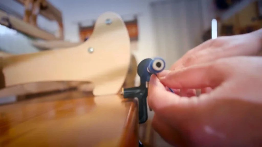 Close-up of a Zoe's hands carefully guiding bright blue yarn onto a bobbin using a manual bobbin winder, with a wooden loom in the blurred background.