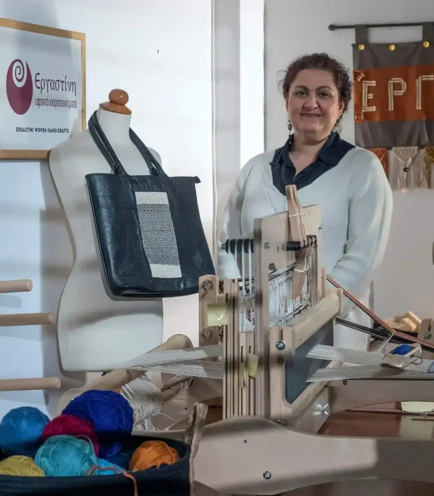 Zoe, founder of Ergastini, in her workshop behind a modern weaving loom, surrounded by colorful yarn and a leather bag with woven embroidery on display.